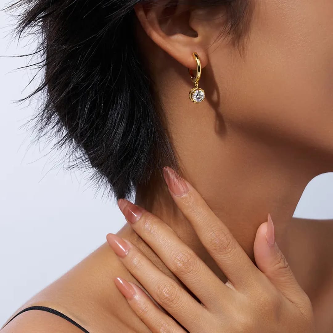 Close-up of a person wearing a gold earring with a clear gemstone on a light background Goldess Jewelley demi fine 18 k gold plated pvd