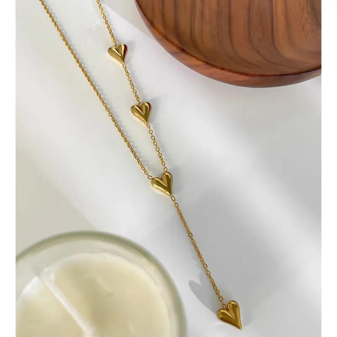 Gold necklace with heart-shaped pendants on a white surface with a wooden bowl in the background. Goldess Jewellry pure 18k gold plated jewellery which last forever