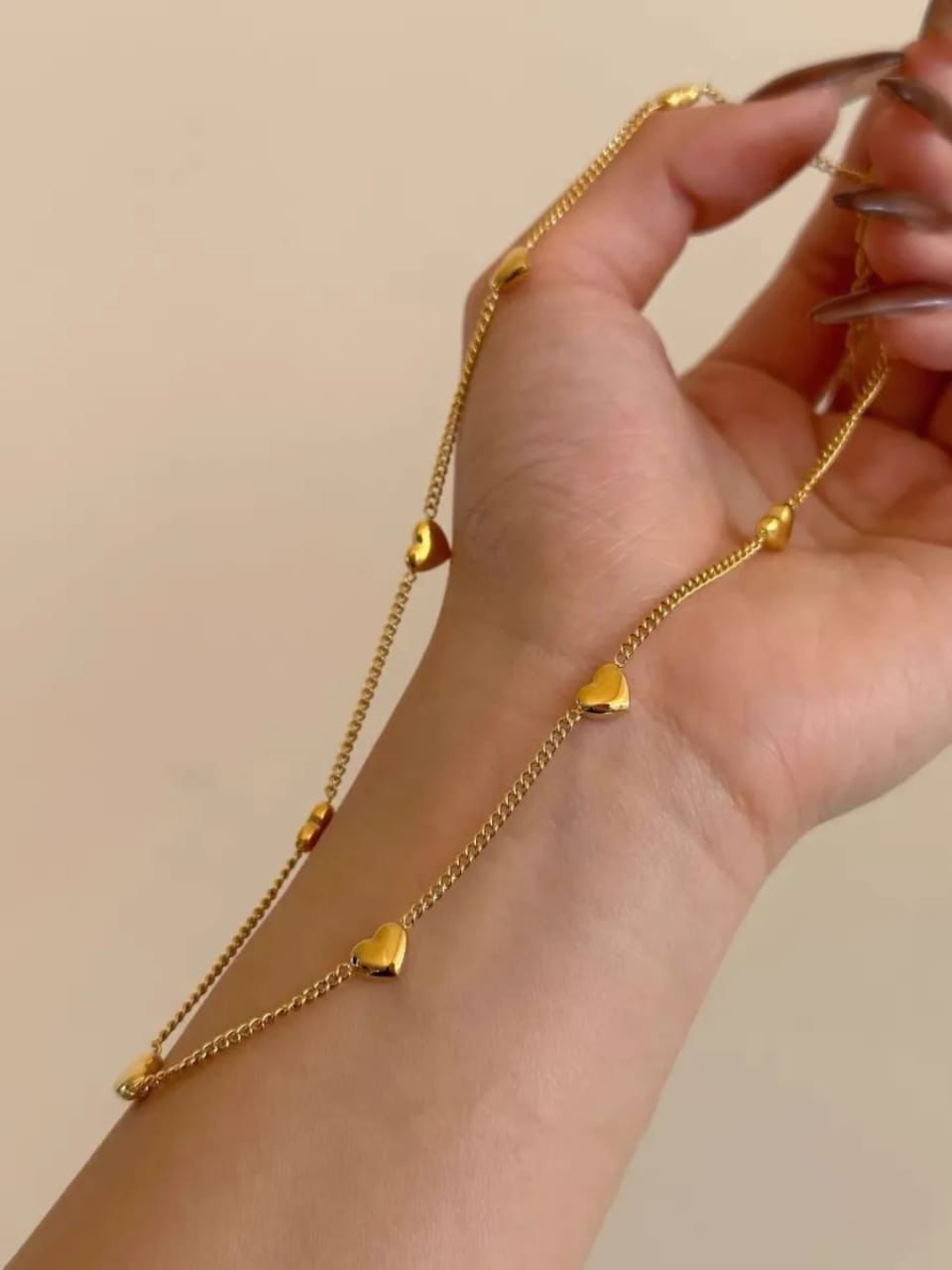 Gold heart-shaped necklace being worn on a wrist against a beige Goldess Jewellry with pvd plating technology which last 10 times than normal jewelley 22background