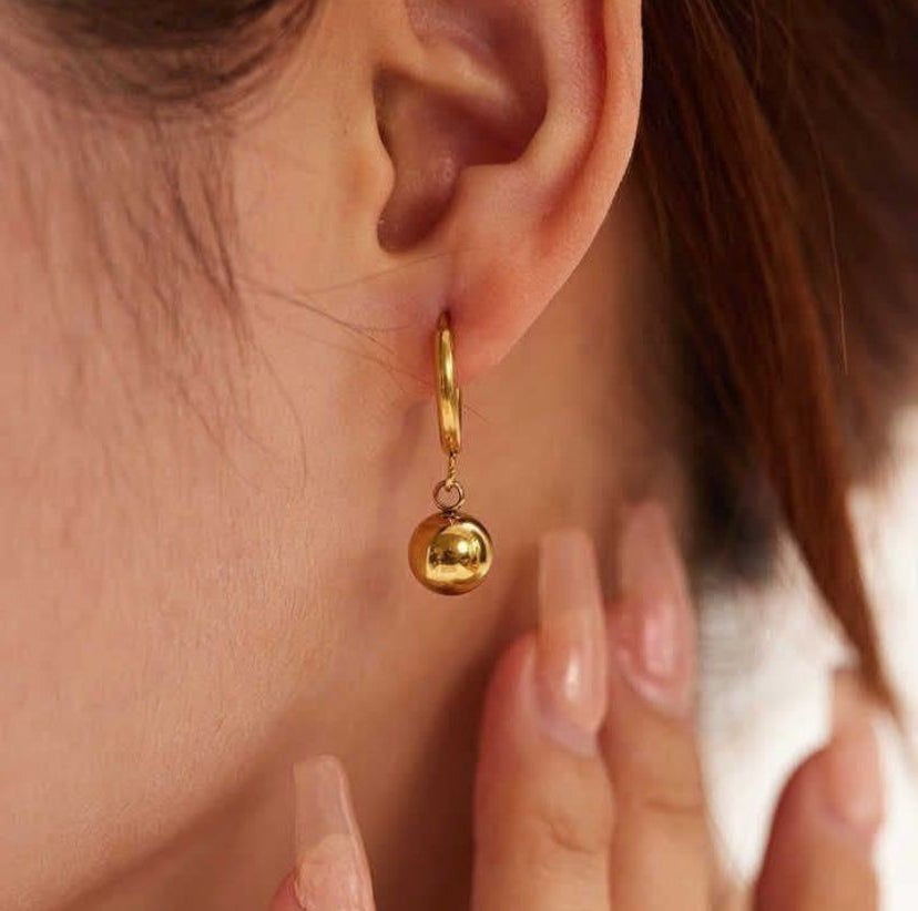 Gold hoop earring with a small gold ball pendant worn by a person.