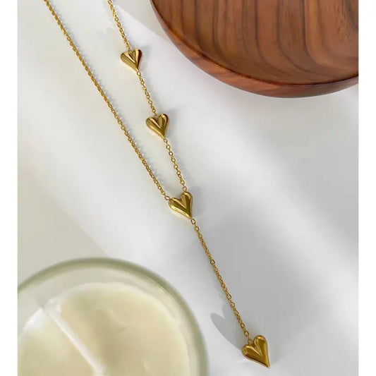 Gold necklace with heart-shaped pendants on a white surface with a wooden bowl in the background. Goldess Jewellry pure 18k gold plated jewellery which last forever