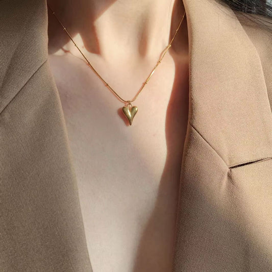 Gold heart-shaped necklace worn by a person in a beige suit. Goldess demi fine jewellery pure 18k gold plated