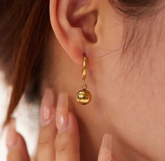 Gold hoop earring with a dangling sphere worn by a person. Goldess Jewellery gold plating pvd technology